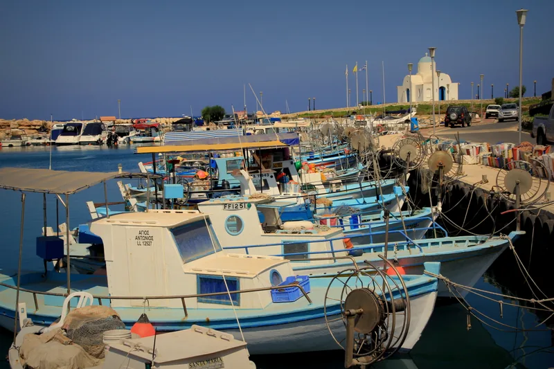Small blue and white fishing boats moored along the quay of Ayia Napa harbour, with a white chapel on the far shore.