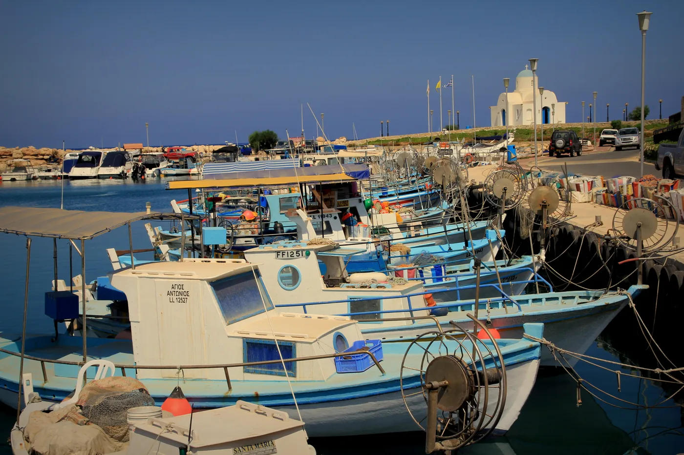 Small blue and white fishing boats moored along the quay of Ayia Napa harbour, with a white chapel on the far shore.