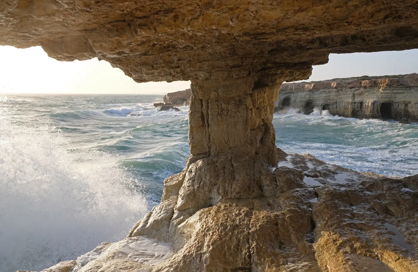 Sea caves at Cape Greco: a pale limestone arch over the water, waves breaking against the cliffs beyond.