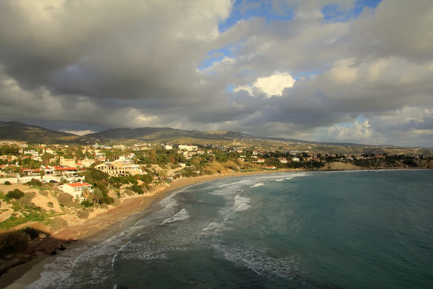 Coral Bay from the headland on a cloudy day: a sandy crescent beach, villas on the slope behind and hills on the horizon.