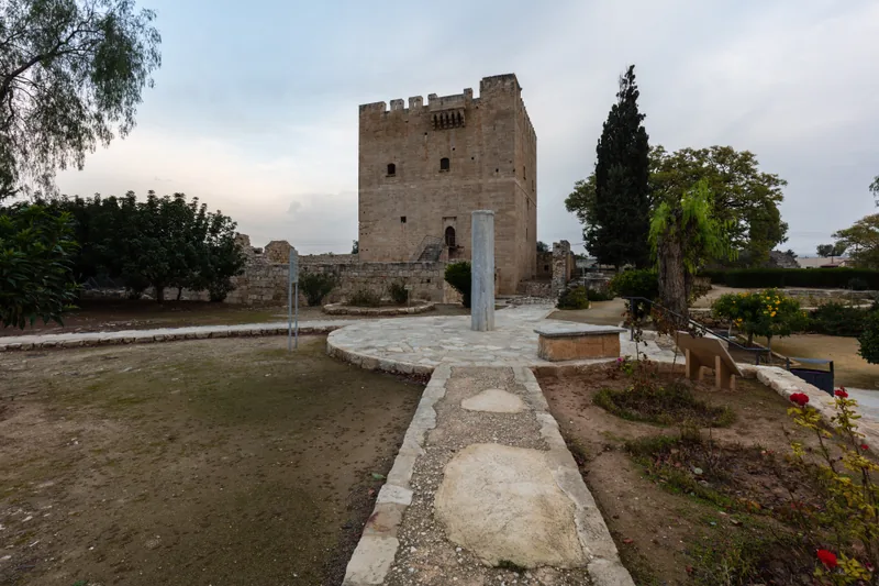 Der quadratische steinerne Bergfried der Burg Kolossi hinter einem gepflasterten Weg und einem Garten, mit hohen Zypressen und Bäumen ringsum.