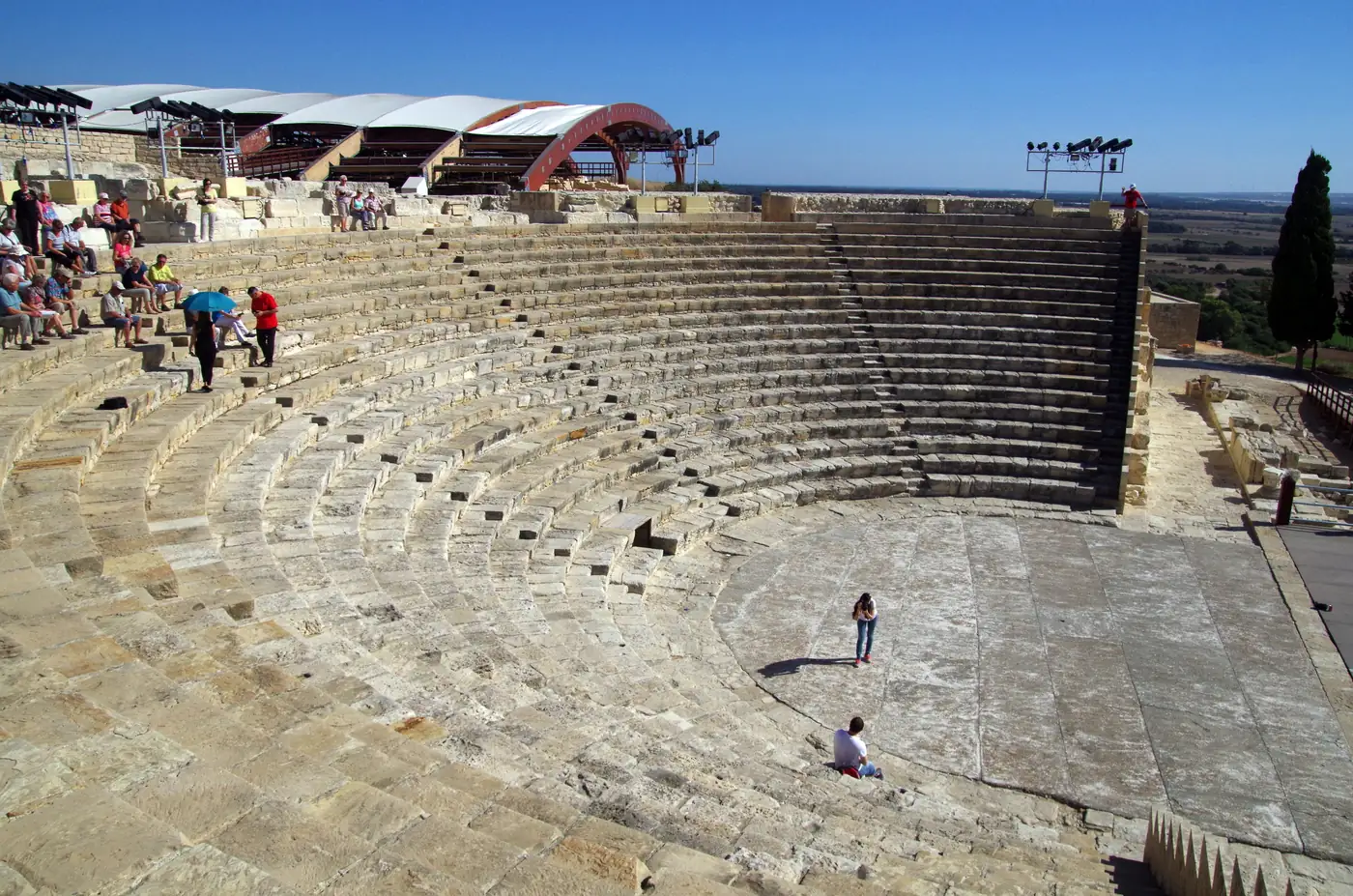 The restored stone theatre at Kourion, a semicircle of seating rows open to the sky, with visitors on the upper tiers and a shade canopy above.