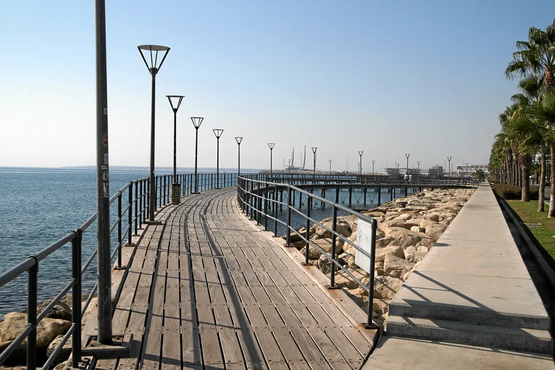 A wooden boardwalk with lamp posts running beside the sea along Limassol’s seafront promenade, palm trees on the right.
