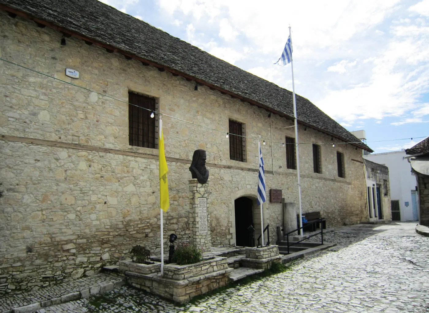 The long stone front of the Holy Cross monastery in Omodos under a stone-slab roof, with a bust on a pedestal and flags by the doorway, beside a cobbled lane.