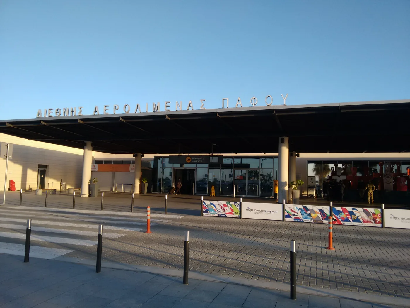 The front of the Paphos airport terminal under a clear sky, its name in large letters along the roof edge, with bollards and an empty forecourt in front.