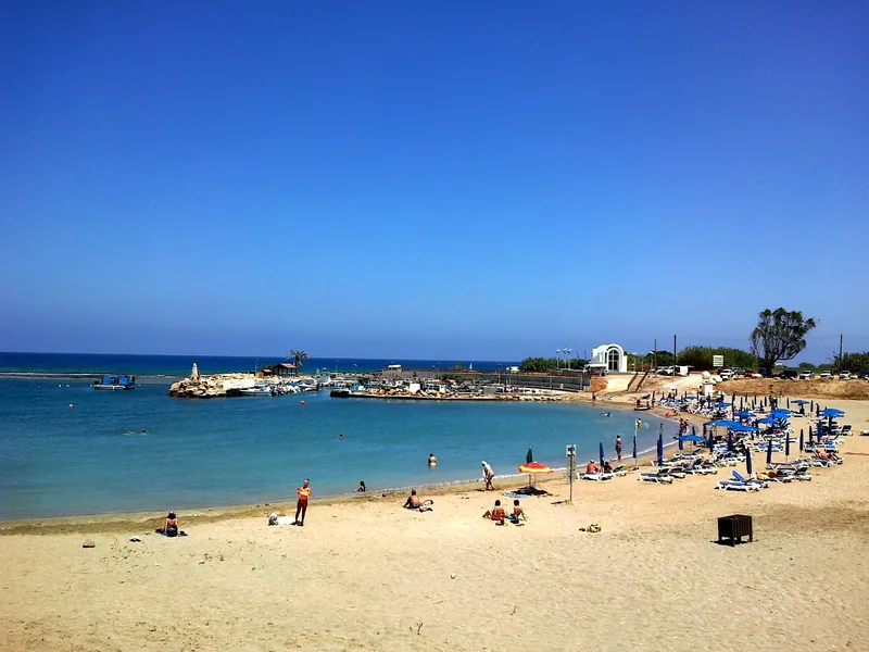 Der Strand Agia Triada bei Paralimni, Sonnenliegen und Schwimmer auf hellem Sand, eine kleine Hafenmauer mit Booten und eine weiße Kapelle dahinter.