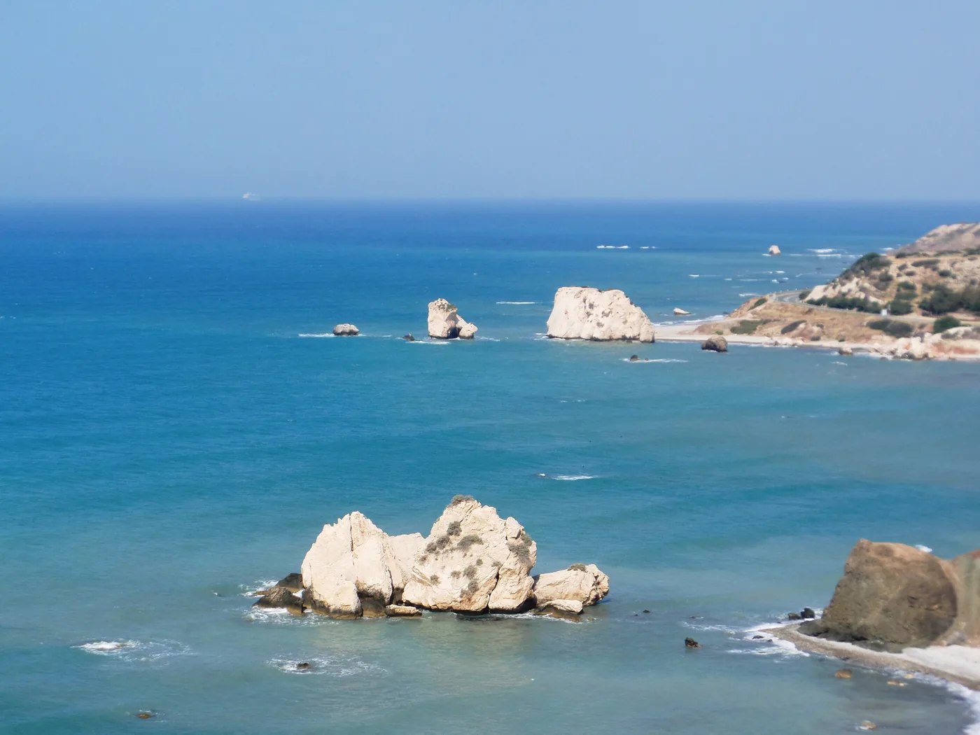 Petra tou Romiou from the cliff: pale limestone sea stacks in clear blue water, a larger rock beyond them and a low headland on the right.