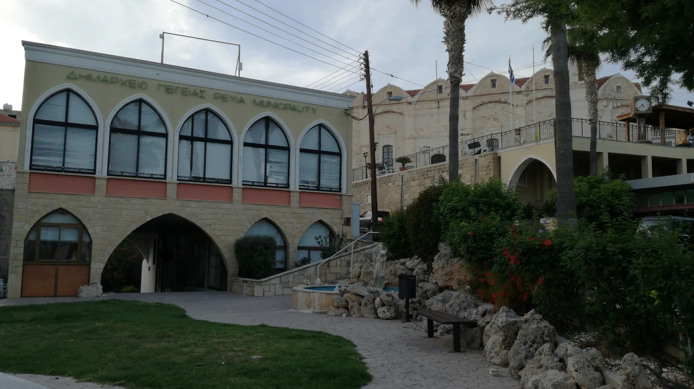 The Peyia municipality building, a pale two-storey front with arched windows and a garden in front, under a cloudy sky.