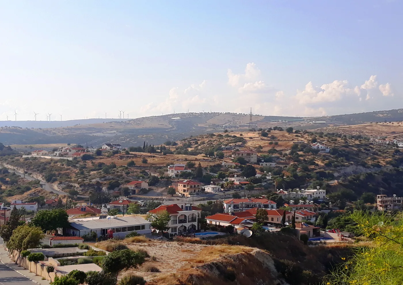 Pissouri village on a hillside, white houses with red roofs among trees, with wind turbines on the ridge behind.