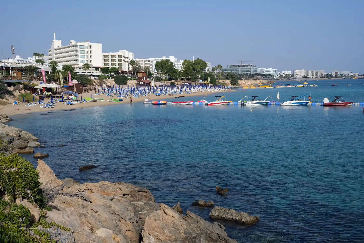 A curve of beach at Protaras seen past rocks in the foreground, with sunshades, small boats on the water and hotels behind.